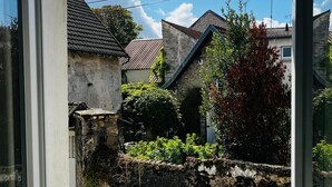 Blick durch ein Fenster auf eine Steinmauer, einen kleinen Garten und ein Haus mit Satteldach.