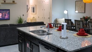 Una cocina moderna con encimeras de granito, un lavamanos doble y lavavajillas. Hay un comedor con mesa y sillas, y una sala de estar con sofá y televisor.