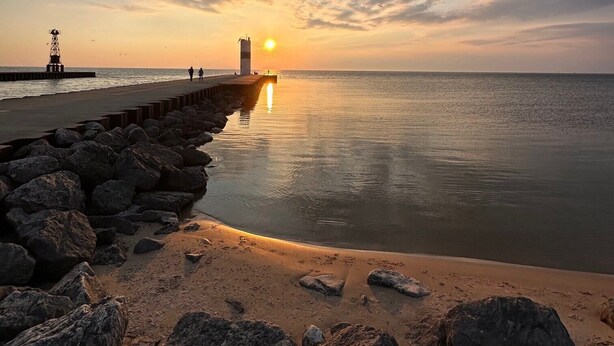 Een kustgezicht met een rotsachtige kustlijn, een pier die het water in loopt en een zonsondergang in de verte.