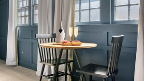 Une pièce avec un mur bleu foncé, une fenêtre ornée de rideaux blancs, une table ronde en bois avec deux chaises, une bouteille de champagne et un verre.