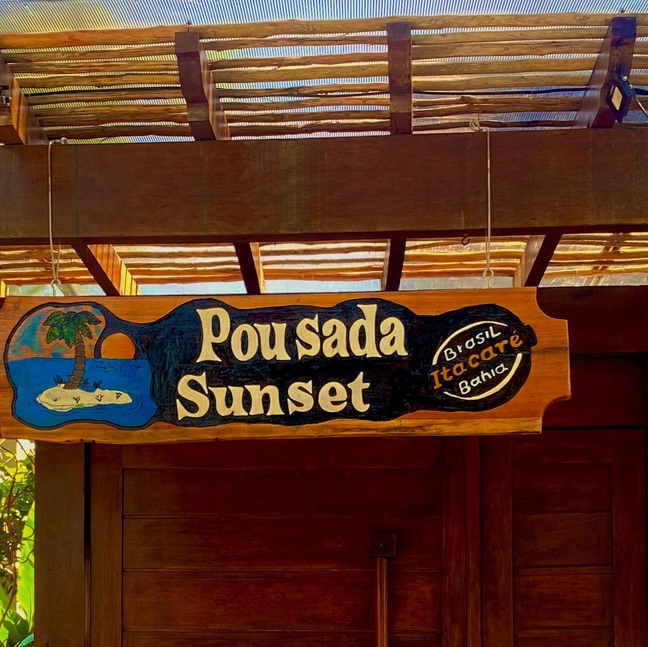 A wooden sign with a palm tree and sunset illustration, hanging from a structure with a roof.