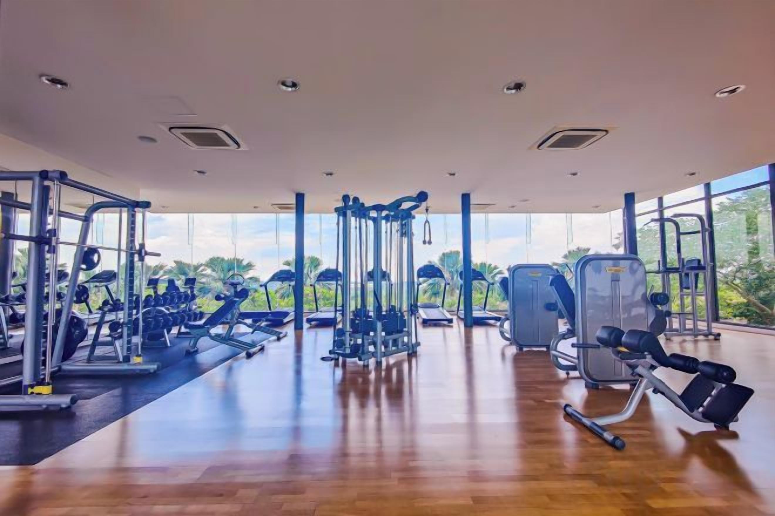 Un gimnasio con diversos equipos de ejercicio, amplias ventanas y suelo de madera.