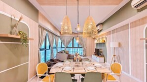 A dining area with a long table set for four, surrounded by chairs with yellow and green cushions. There are hanging wicker pendant lights above the table, and the room has large windows with curtains.