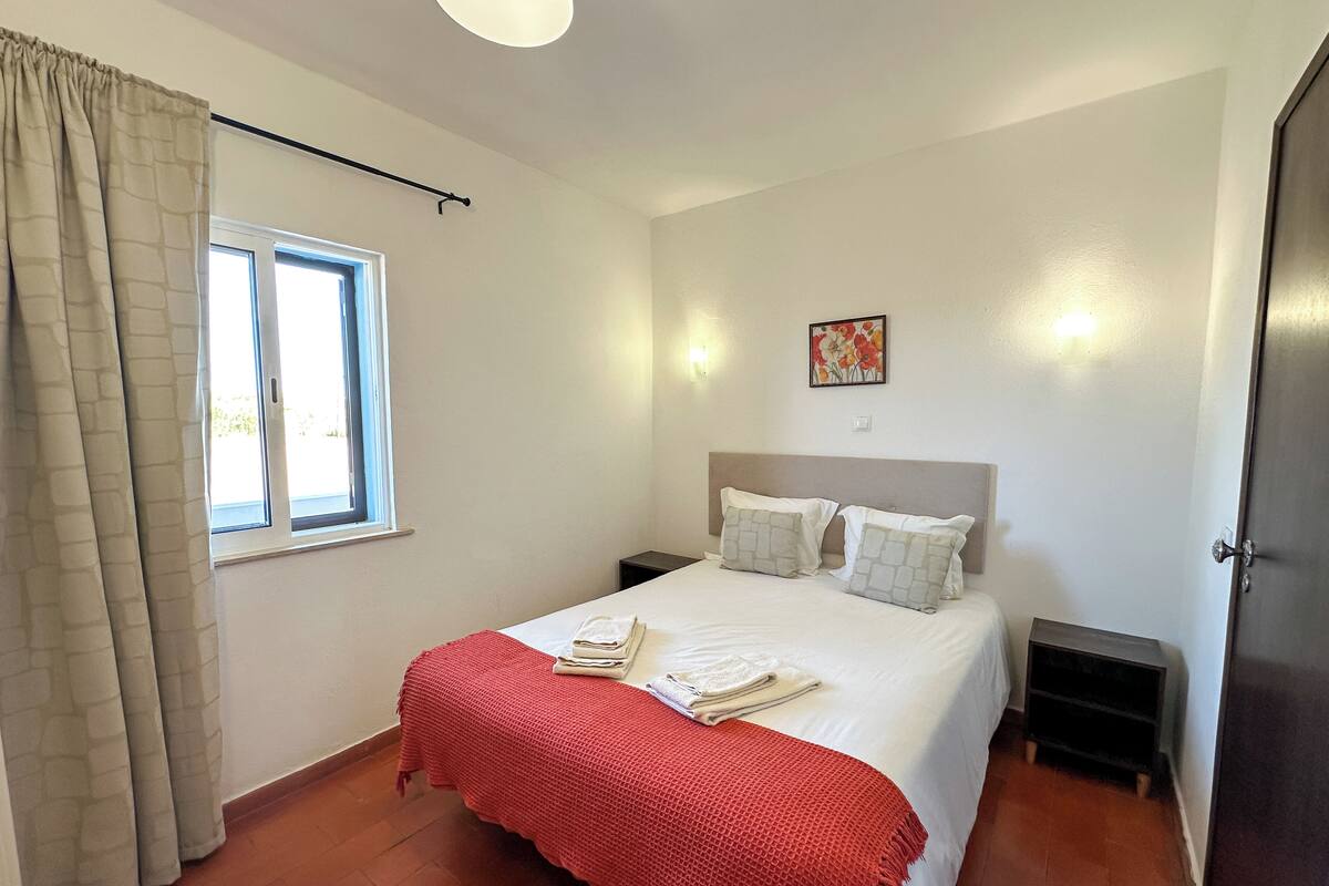 Un dormitorio con una cama, dos mesitas de noche, una ventana con cortinas, una lámpara de pared y una puerta.