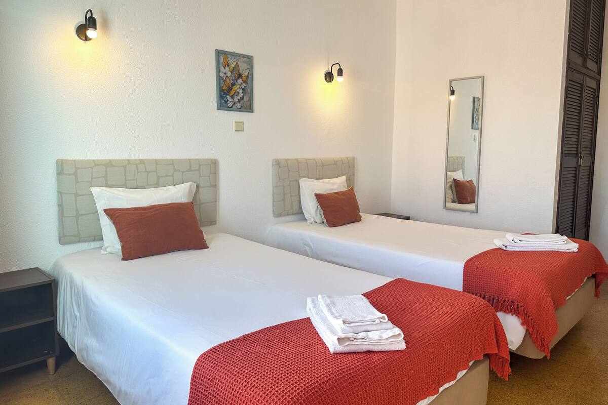Habitación de hotel con dos camas, cada una con cabecero, ropa de cama blanca y una manta roja. Hay luminarias fijadas a la pared y un cuadro sobre las camas.