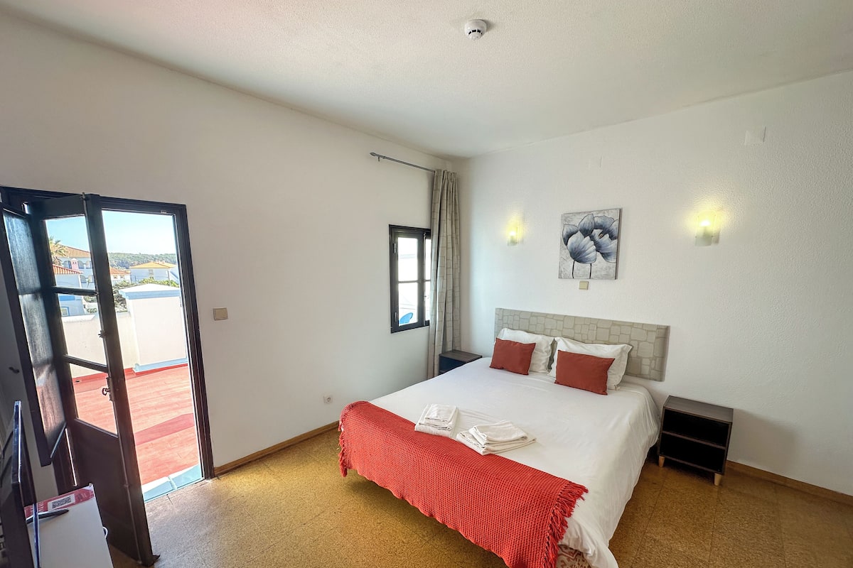Habitación de hotel con cama, ventana, puerta y vistas a edificios.