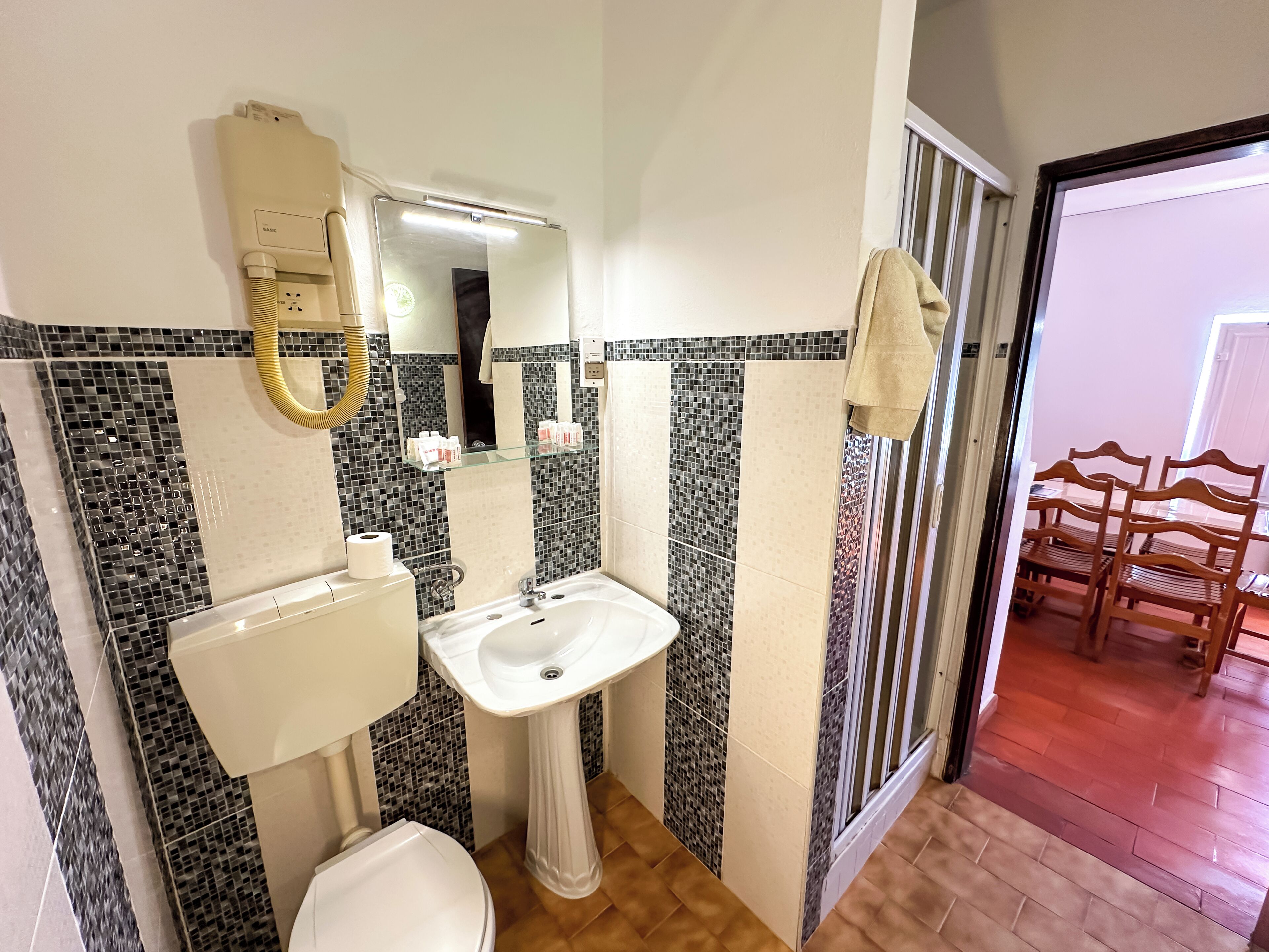 A bathroom with a toilet, sink, and mirror. There is a towel hanging on the wall and a chair in the adjacent room.