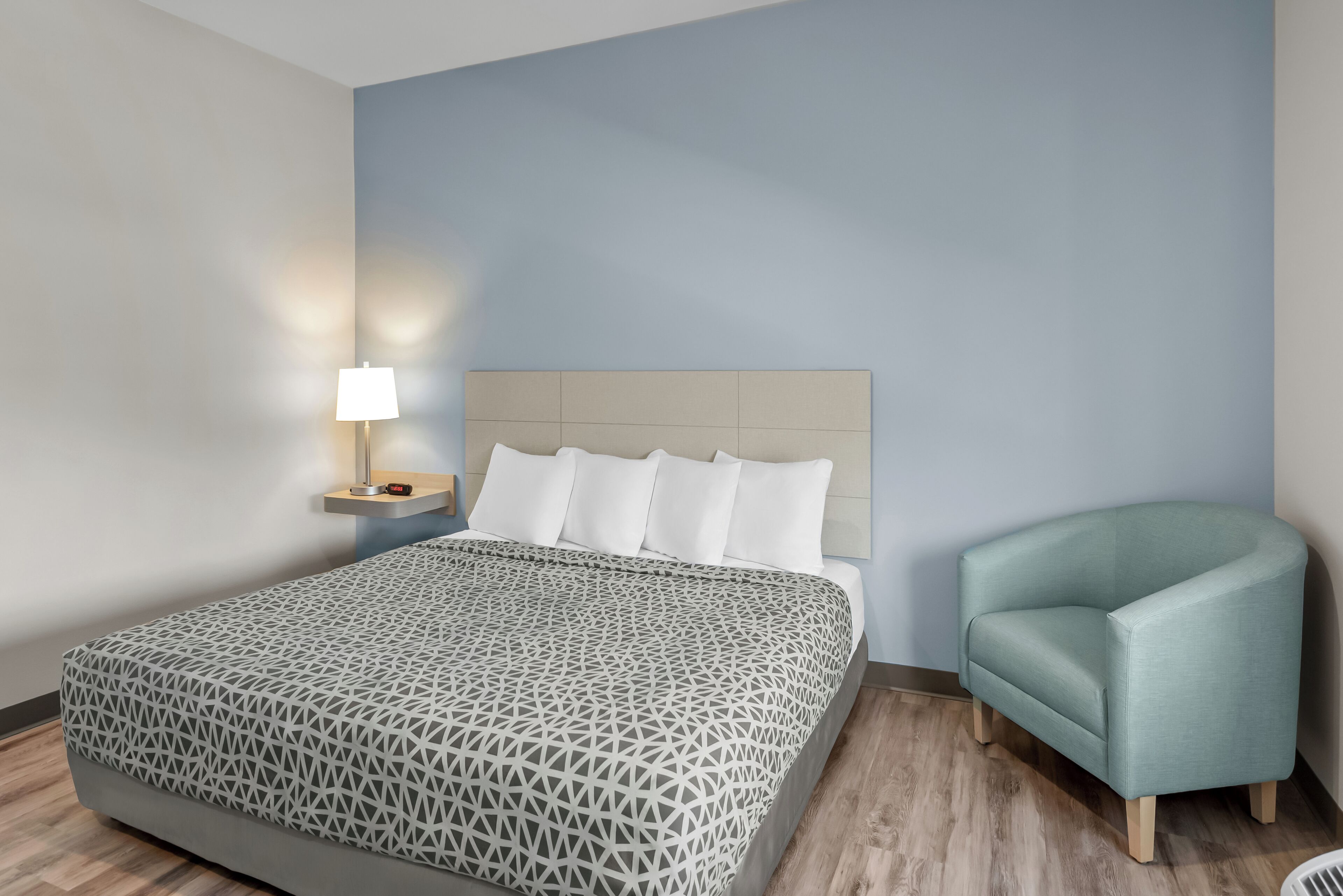 A bedroom with a bed, a nightstand with a lamp, and a light blue armchair.