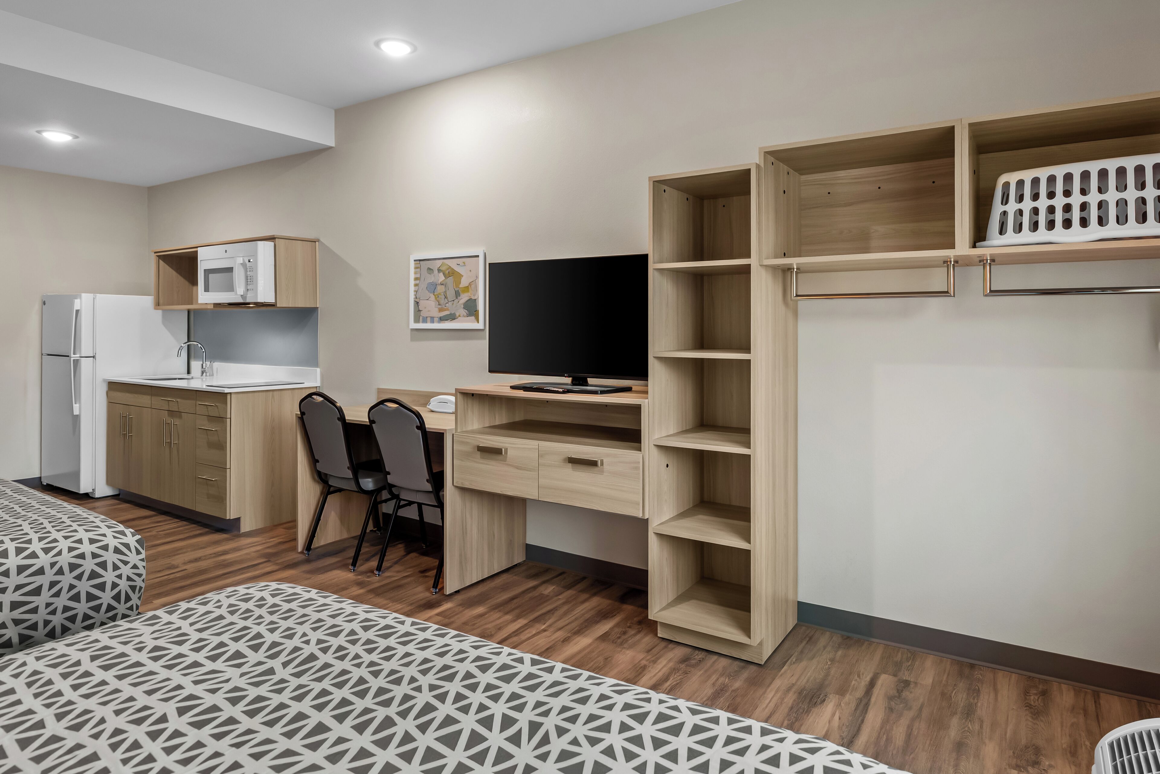 A compact room with a kitchenette, a desk with a chair, and two beds with patterned bedding.