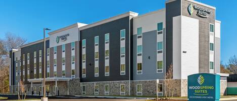 Un hôtel à plusieurs étages au design moderne, doté d’un logo bien visible et d’une signalétique indiquant qu’il s’agit d’un hôtel Woodspring Suites proposant des séjours de longue durée.