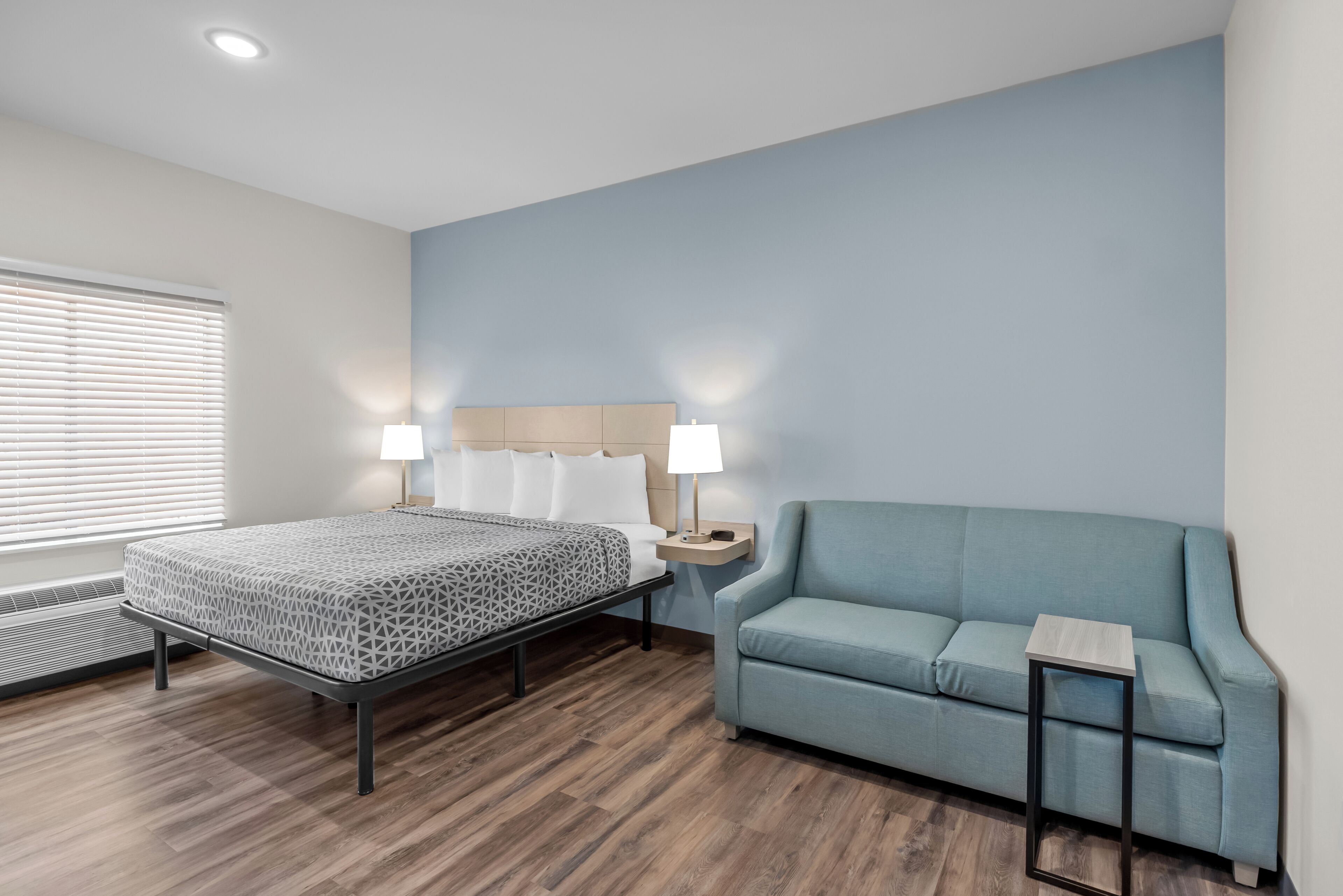 Een hotelkamer met een bed, een slaapbank, een nachtkastje met lamp en een raam met jaloezieën.