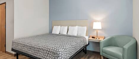 Una cama bien hecha con una colcha estampada, una lámpara de noche y un sillón verde en una habitación con suelo de madera y paredes azul claro.