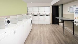 A laundry facility with multiple washing machines and dryers, a table, and a door. - WoodSpring Suites Cleveland North (Cleveland)