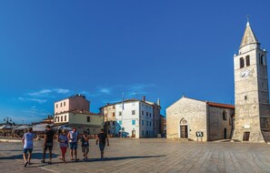 A square with stone buildings, a church with a clock tower, and people walking.