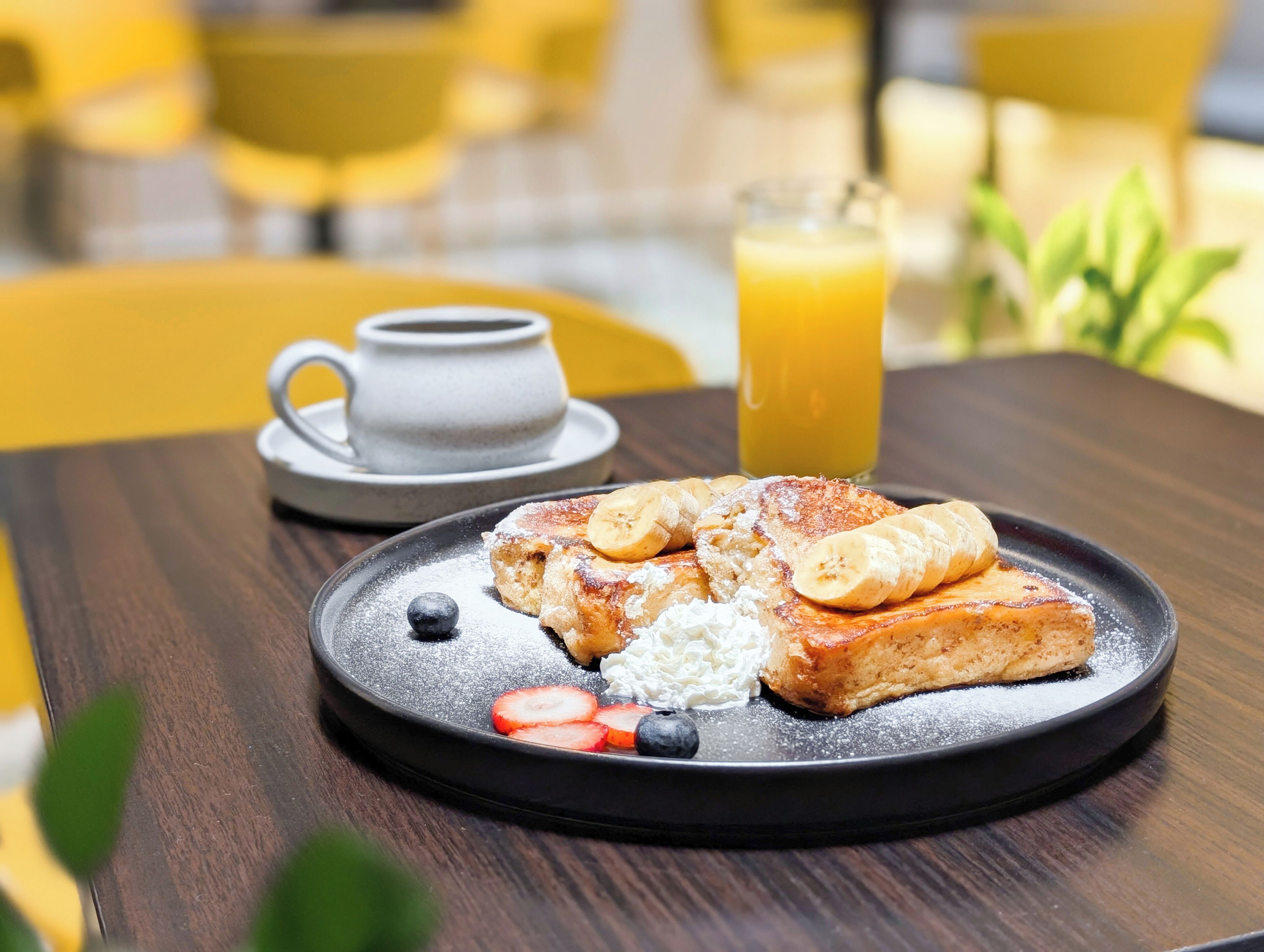 A breakfast plate with pancakes, bananas, strawberries, and blueberries, a cup of coffee, and a glass of orange juice on a wooden table.