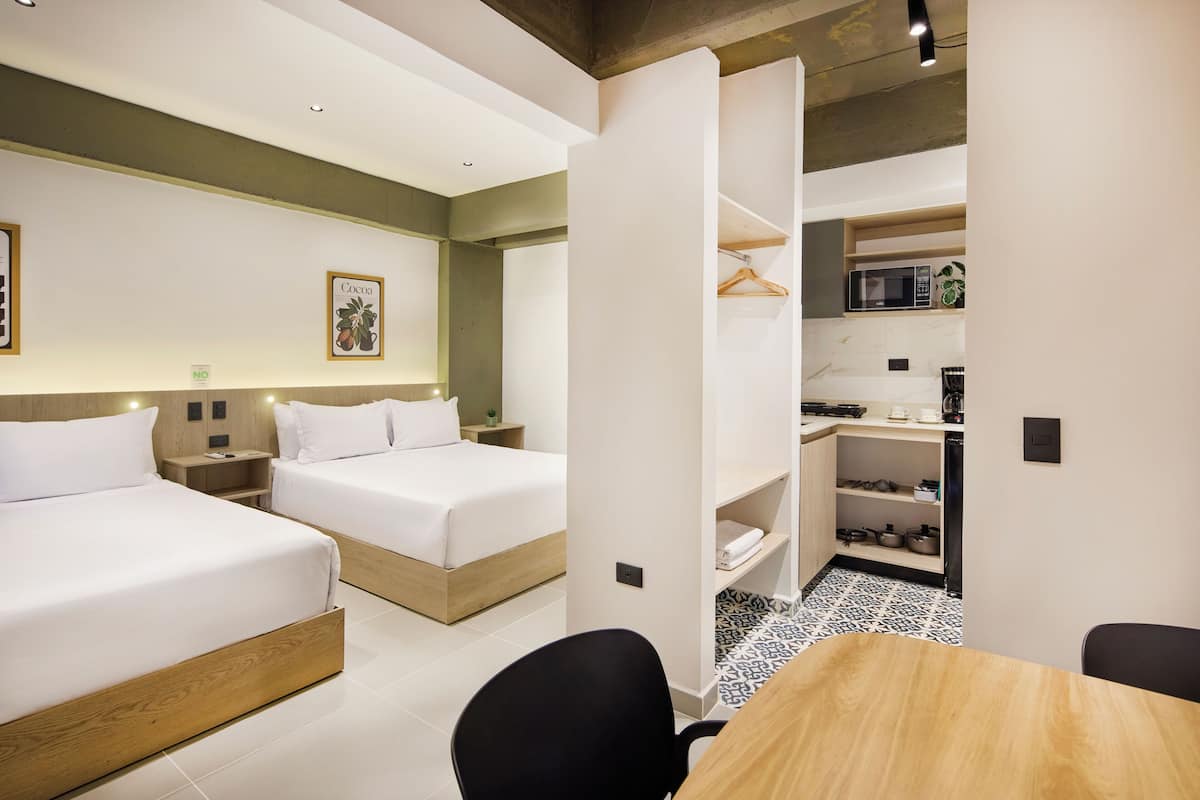 A modern hotel room with two beds, a small kitchenette, and a dining area.