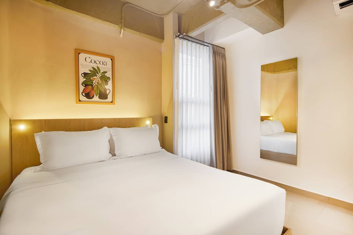 A hotel room with a large bed, a framed picture of cocoa, a mirror, and a window with curtains.
