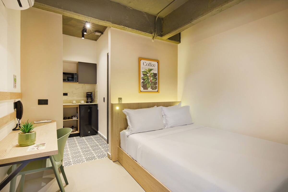 A compact hotel room with a bed, a desk with a plant, a microwave, and a small kitchenette.