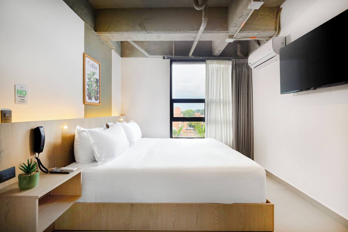 A modern hotel room with a large bed, a mounted TV, a phone on the nightstand, and a view of the city through the window.