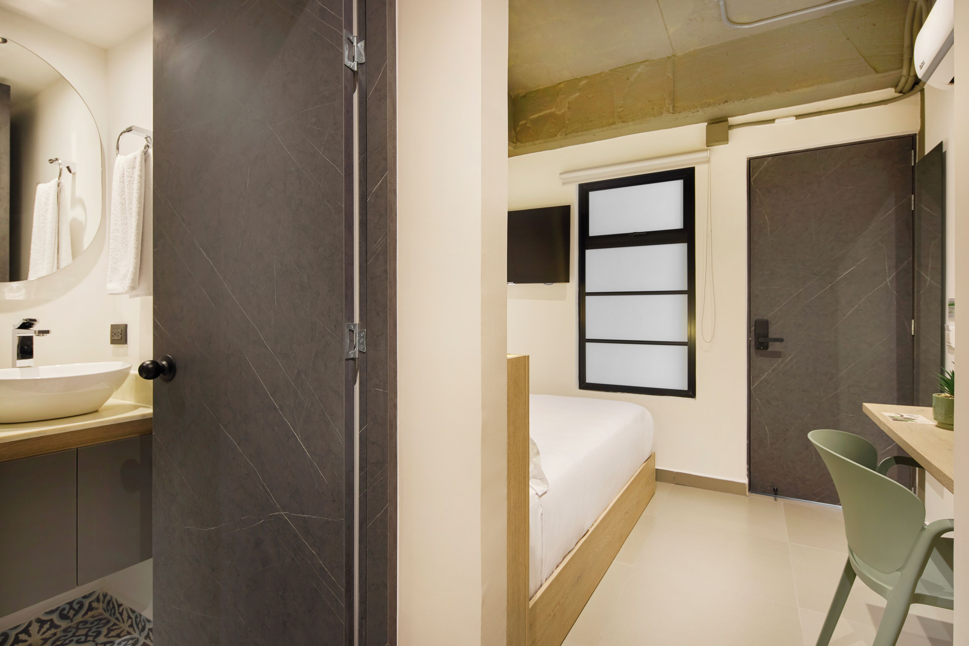 A modern bedroom with a bed, a bedside table, a chair, a window, and a bathroom with a sink and mirror.