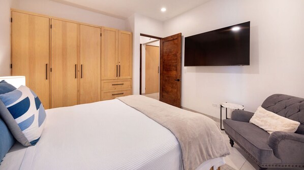Un dormitorio con cama, sofá gris, armario de madera y televisor montado en la pared.