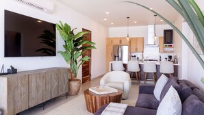A modern living room with a large TV, a potted plant, a sofa, and a dining area with bar stools. - Oceanfront 2BR • Elevated Views • Large Terrace (Los Cerritos)
