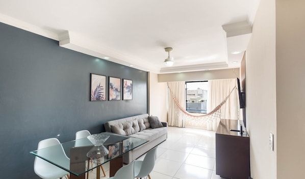 A modern living room with a glass dining table, grey sofa, and a hammock by the window.