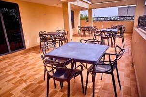 Un área de comedor al aire libre con techo, con varias mesas y sillas, suelo de baldosas y vistas del entorno.
