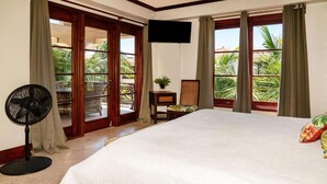 A hotel room with a bed, a TV, a fan, a small table with a plant, and a view of palm trees through the window. - La Beliza Resort (San Pedro)