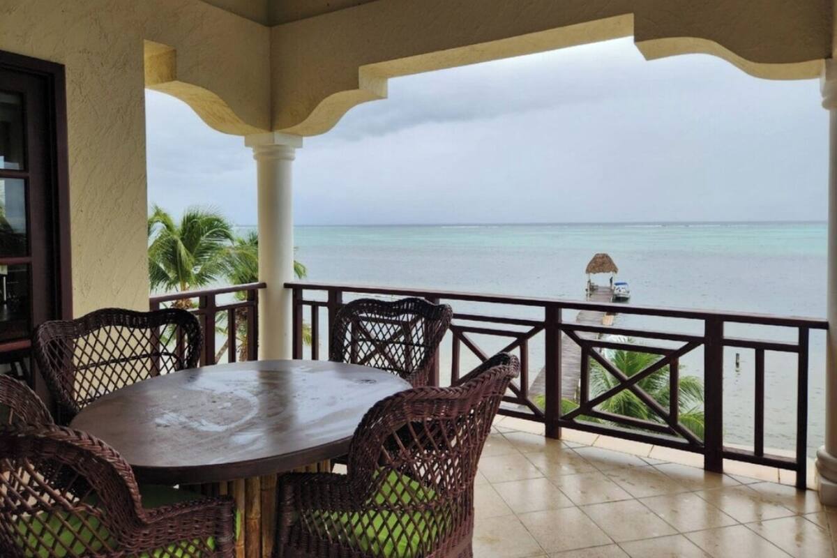Un espacio de estar techado al aire libre con una mesa y sillas de mimbre, con vista a una playa con un muelle y un bote.