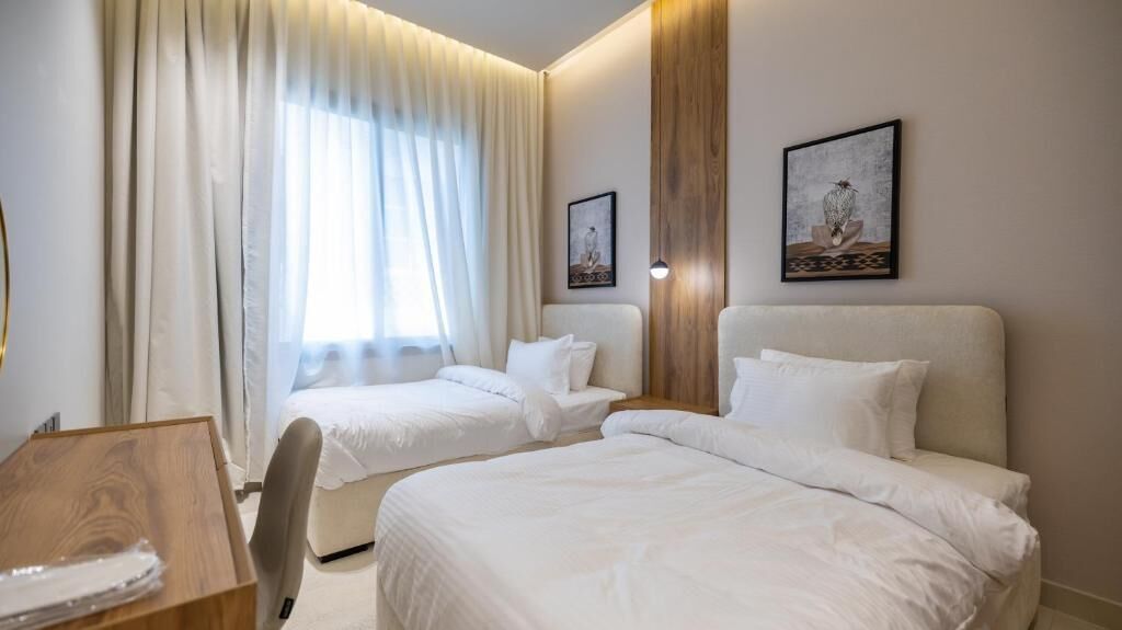 Habitación de hotel con dos camas, un escritorio y una silla. Cortinas en la ventana y dos cuadros en la pared.