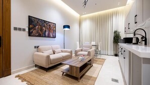 A modern living room with a sofa, armchairs, and a coffee table. There is a painting on the wall, a floor lamp, and a kitchen area with a sink and cabinets. - Super Luxury two Bedroom Al Narjis (Riyadh)