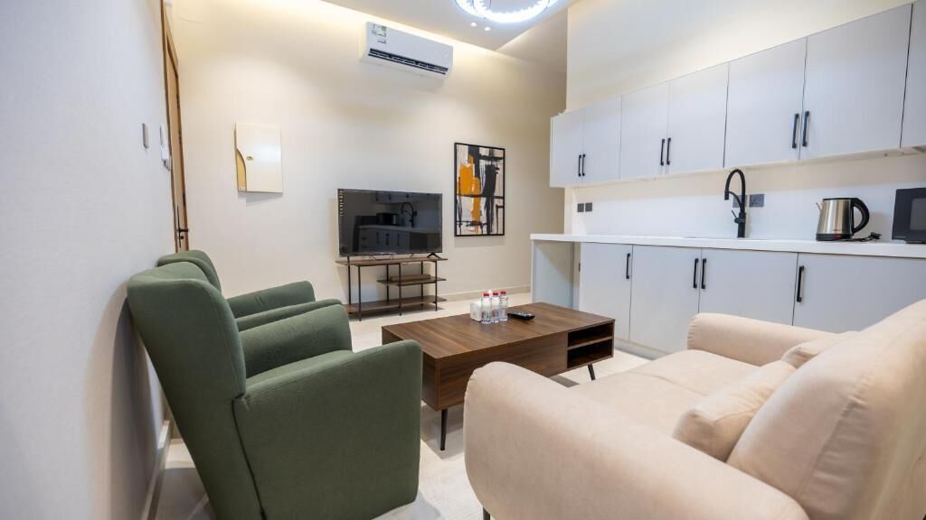 A modern living room with a sofa, a coffee table, and a TV. There is a kitchen area with cabinets and a microwave.