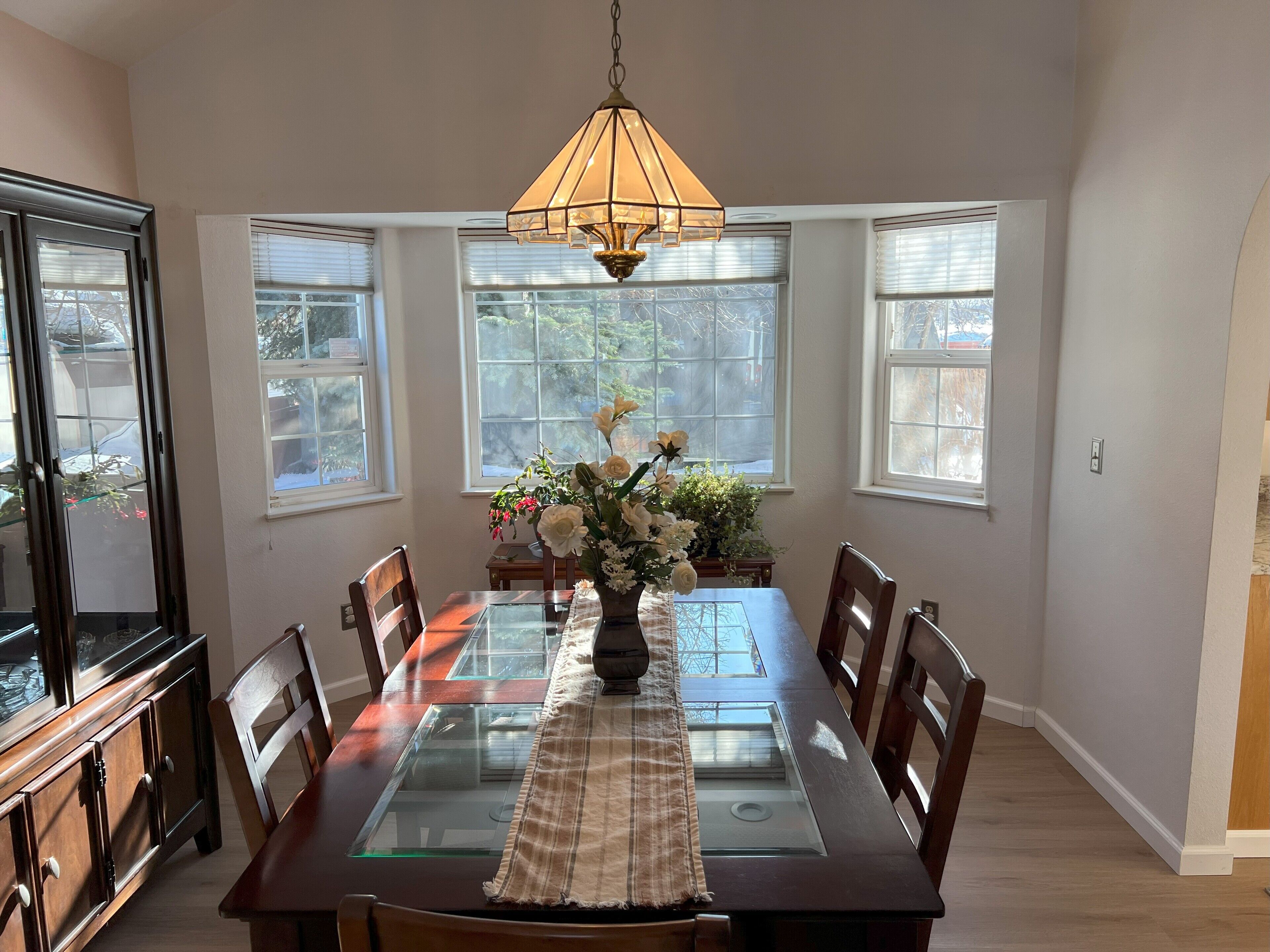 Comedor con mesa de vidrio, sillas de madera, un jarrón de flores y una lámpara colgante.