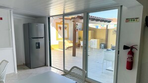 A compact kitchen with a refrigerator, a dining table with chairs, and a sliding glass door leading to an outdoor patio area. - Casa Alagoinha by Umbral (Odemira)