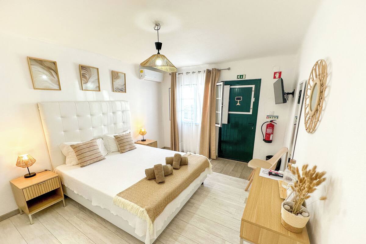 Habitación de hotel con una cama grande, mesitas de noche, un escritorio, una lámpara colgante, un extintor y una puerta verde.
