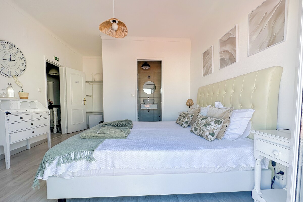 A spacious bedroom with a large bed, a bedside table, a wardrobe, and a bathroom visible through an open door.