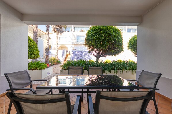 A balcony with a glass table and four chairs, overlooking a landscaped garden and a building with balconies.