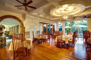 A spacious dining area with wooden tables and chairs, large windows offering a view of greenery, and a ceiling fan.