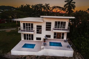 Uma casa moderna de dois andares com telhado plano, grandes janelas e área de piscina central com escadas que levam a um pátio.
