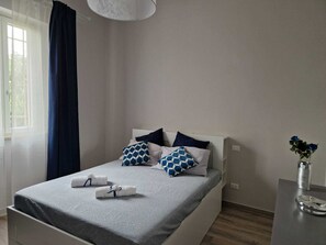 Een net opgemaakt bed met een grijze dekbedovertrek, twee blauwe kussens met een patroon, en twee opgerolde handdoeken.