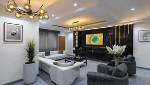 A modern living room with a sofa set, a coffee table, and a grey armchair. There is a TV mounted on the wall and a plant in a white pot. - Daveange Hotels (Port Harcourt)