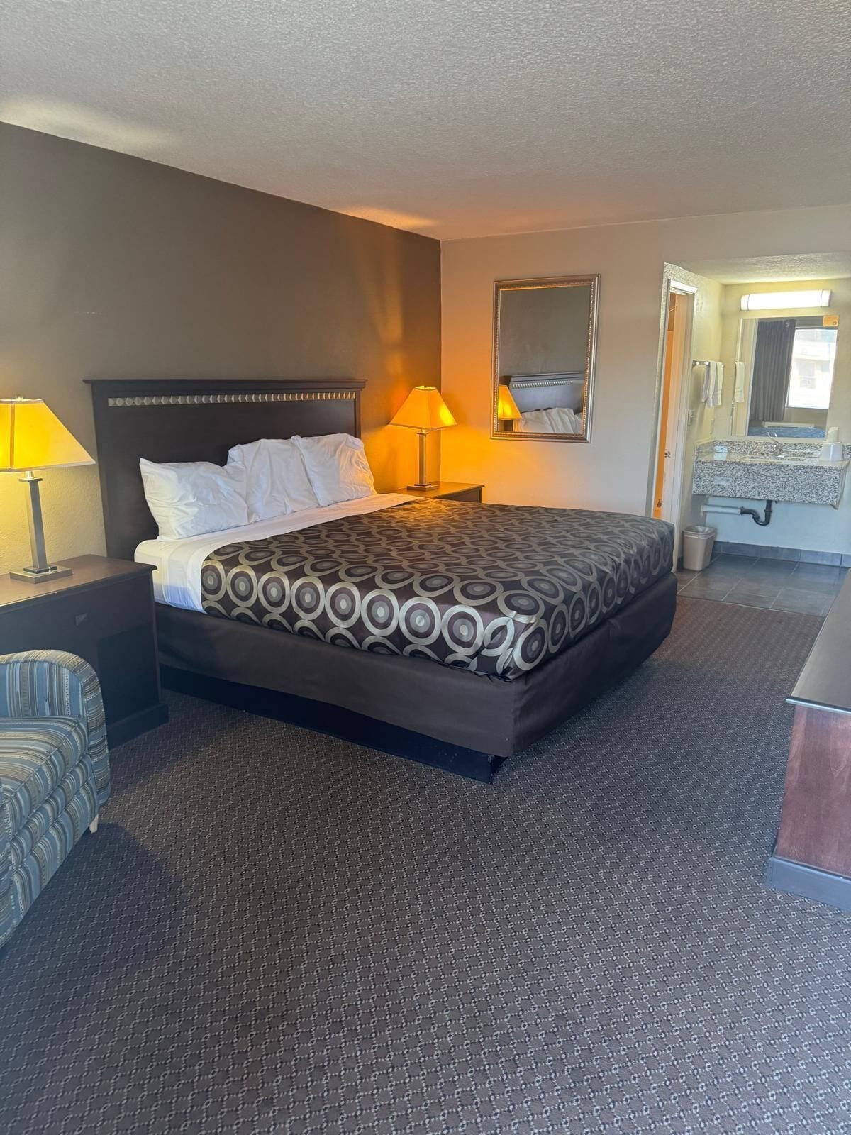 Une chambre d’hôtel avec un lit, deux lampes de chevet, un miroir, une petite table, une chaise et une salle de bain visible à travers une porte ouverte.