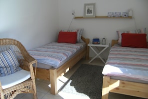 A bedroom with two single beds, a wicker chair, a small table, and a shelf with pictures.