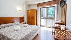 Een hotelkamer met een groot bed, een houten garderobe, een bureau met stoel, een aan de muur bevestigde televisie en een glazen schuifdeur naar een balkon.