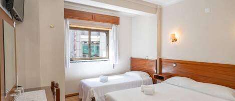 A hotel room with two single beds, a wooden headboard, a desk with a chair, a TV mounted on the wall, and a window with curtains.