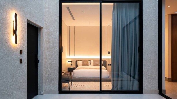 A modern hotel room with a large sliding glass door, a bed with white linens, a nightstand, and a wall-mounted light fixture. - Nolina chalets (Madinah)
