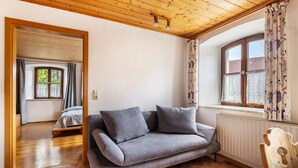Ein Zimmer mit Holzboden, einem grauen Sofa, einem Holzstuhl, einem Fenster mit gemusterten Vorhängen und einem Heizkörper.