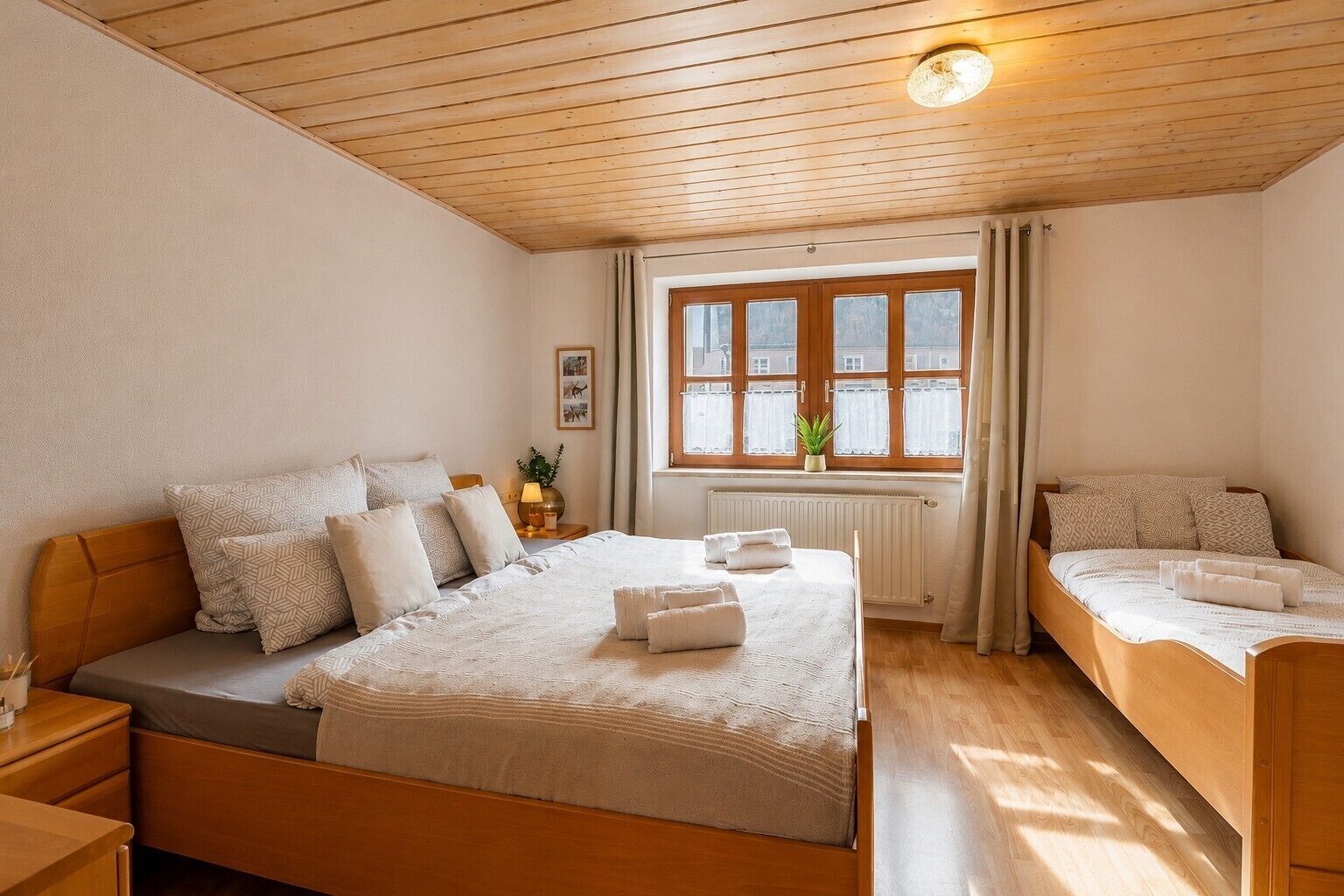 Un dormitorio con una cama de madera, una cama más pequeña, una mesita de noche, una ventana con cortinas y un radiador.