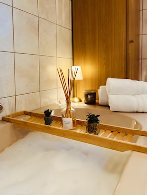 Une salle de bain avec un plateau en bois contenant un flacon de diffuseur à bâtonnets, deux petites plantes en pot et une serviette blanche posée sur la baignoire.
